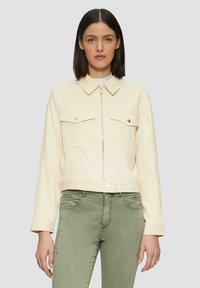 Vrouw met recht zwart haar die een crèmekleurige ritsjack met knoopzakken op de borst en olijfgroene broek draagt, tegen een effen achtergrond.