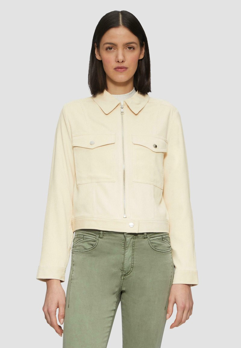 Vrouw met recht zwart haar die een crèmekleurige ritsjack met knoopzakken op de borst en olijfgroene broek draagt, tegen een effen achtergrond.