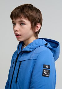 Chaqueta azul con capucha, que presenta un tejido texturizado, cremallera negra y bolsillos laterales. Un parche de logo negro se encuentra en la manga izquierda.