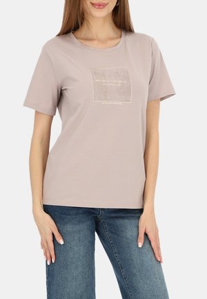 Kobieta w beżowym t-shircie z krótkim rękawem z małym napisem i grafiką na piersi, w połączeniu z niebieskimi dżinsami, stoi z rękami spoczywającymi po bokach.