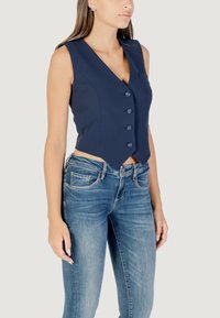 Gilet sans manches bleu marine avec un col en V et une fermeture à cinq boutons, présentant une coupe ajustée et un détail de poche subtil. Associé à un jean bleu.