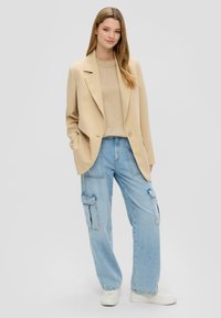 Beige oversized blazer, lichtbruine top en losse lichtblauwe cargobroek met zijzakken, gecombineerd met witte sneakers. Eenvoudige, relaxte pasvorm.