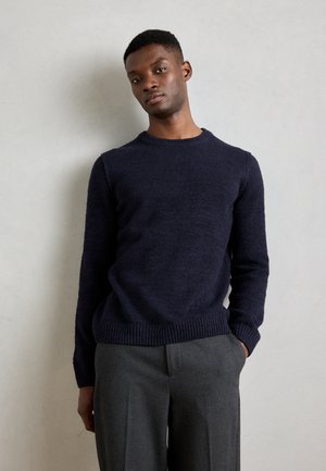 Pull en maille bleu marine avec un col rond, des poignets et un ourlet côtelés. Associé à un pantalon large gris, présentant une texture intéressante.