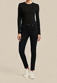 Woman wearing a black long-sleeve top, black high-waist skinny jeans, and white sneakers against a plain backdrop.