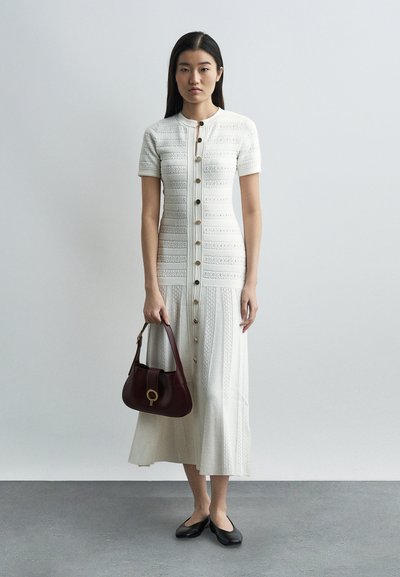 Γυναίκα που φοράει λευκό πλεκτό μίντι φόρεμα με κουμπιά, κρατώντας μια σκούρα καφέ τσάντα, στέκεται σε γκρίζο πάτωμα μπροστά σε απλό ανοιχτό γκρι τοίχο.