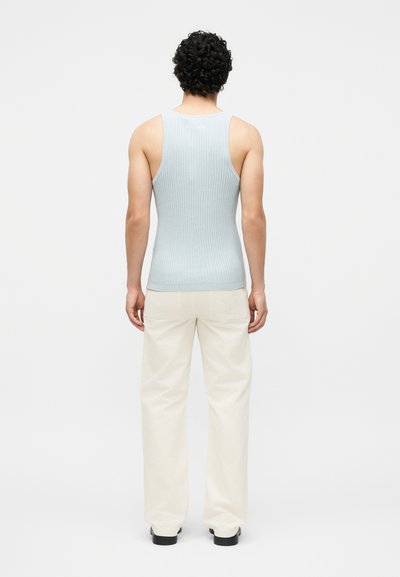 Άνδρας με σγουρά μαλλιά, φοράει ανοιχτό μπλε ριπ αμάνικη μπλούζα, κρεμ παντελόνι και μαύρα παπούτσια, στέκεται αντικριστά σε απλό λευκό φόντο.