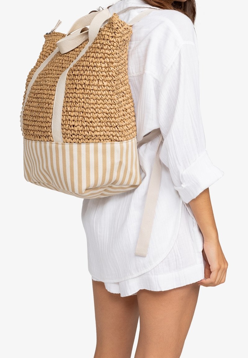 Femme portant une robe chemise blanche avec un sac à dos tissé beige, fond en tissu rayé et bretelles en tissu sur le dos.