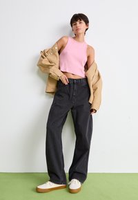Top court rose côtelé, veste beige oversize, jeans noirs à jambes larges et baskets crème avec semelles marron sur un sol vert.