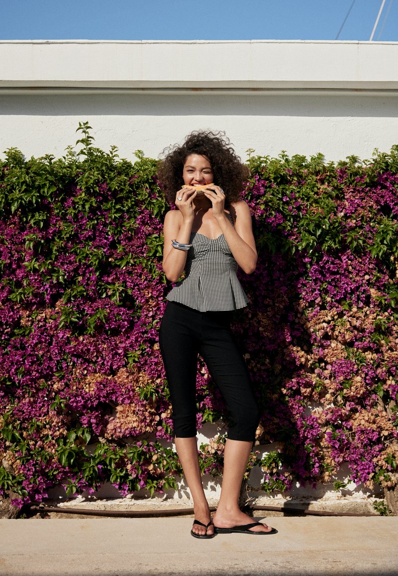 Femme aux cheveux bouclés portant un haut noir et blanc et un pantalon noir, mangeant un sandwich, debout devant un mur avec des fleurs violettes.