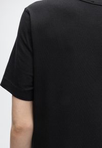 Κοντινό πλάνο ενός μαύρου T-shirt με κοντά μανίκια που φοριέται στην πάνω πλάτη και το χέρι ενός ατόμου, δείχνοντας την υφή του υφάσματος και τις λεπτομέρειες της ραφής.