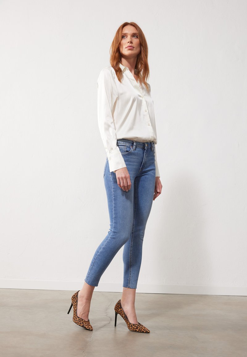 Blusa di seta bianca con bottoni, jeans azzurri aderenti e décolleté a punta con stampa leopardata. La modella è in piedi contro un muro neutro.