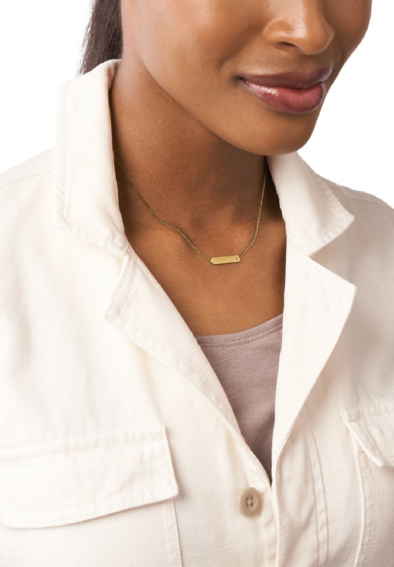 Collana d'oro con un pendente rettangolare, caratterizzata da una catena delicata. Indossata sopra una camicia beige chiaro e un top lilla morbido.