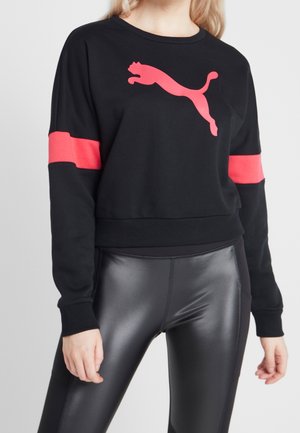 Kvinde iført en sort sweatshirt med rød Puma-logo og røde striber på ærmerne, kombineret med skinnende sorte leggings.