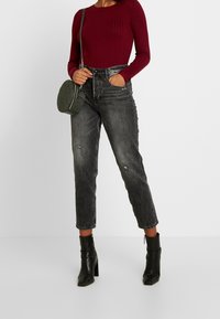 Top long à manches longues bordeaux côtelé, jeans noirs taille haute avec des détails déchirés, associé à un sac circulaire vert et des bottines noires.