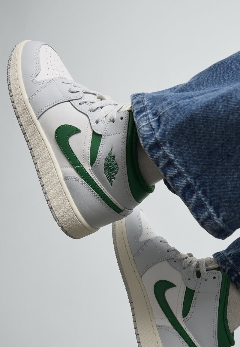 Baskets montantes blanches et vertes portées avec un jean bleu, vues de profil avec les pieds levés sur un fond clair uni.