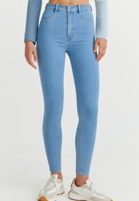 Vaqueros de denim azul de cintura alta con un corte ajustado, con cierre de botón, cinco bolsillos y una textura suave. Llevados con zapatillas blancas y beige.