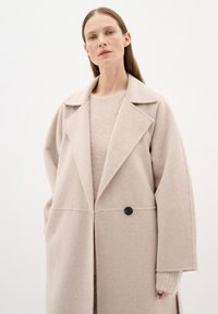 Cappotto in lana beige con ampi revers, maniche lunghe e un solo bottone nero. Indossato sopra un pullover lavorato a maglia beige chiaro con una finitura testurizzata.