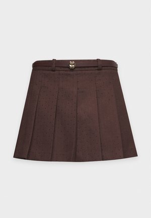 Un mannequin porte un pull à losanges vert, bleu marine et blanc, une minijupe plissée marron, des chaussettes blanches et des chaussures à talons marron, et tient un sac à main beige à franges.
