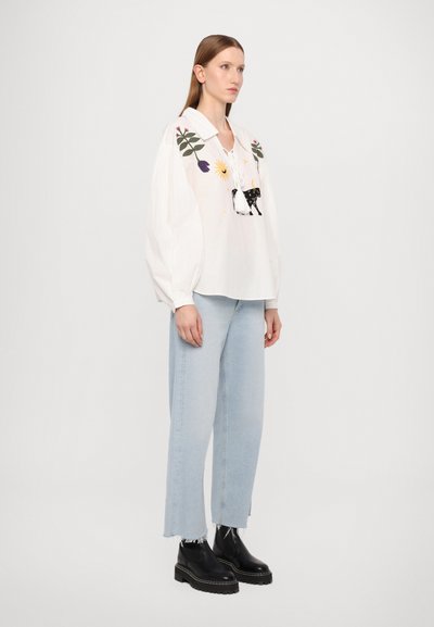 Weiße Bluse mit langen Ärmeln, floralem Stickmuster und verstellbarem Ausschnitt. Kombiniert mit hellblauen gerade geschnittenen Jeans und schwarzen klobigen Stiefeln.