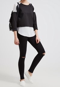 Sudadera de bloque de color en blanco y negro con un cuello redondo, jeans ajustados negros rasgados y zapatillas blancas sin cordones. Incluye una mochila negra con flecos.