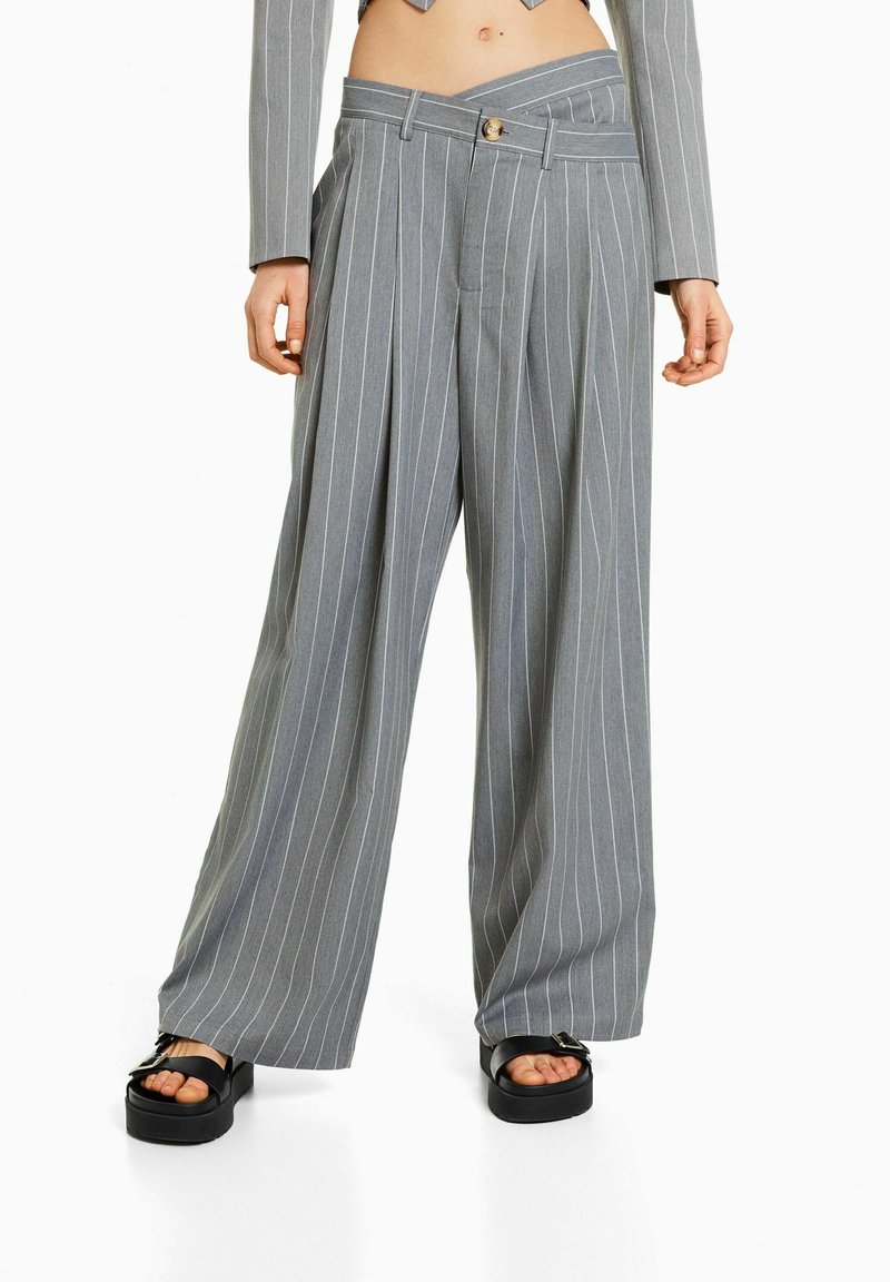 Pantalon large gris à fines rayures avec ceinture asymétrique, porté avec des sandales à plateforme noires, manches courtes de blazer gris visibles.