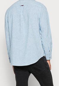 Vaaleansininen ja valkoinen raidallinen pitkähihainen paita, jossa on nappimanchetit ja suora helma. Selkäpuolella on laskos ja logotyyppitys kaulan lähellä.