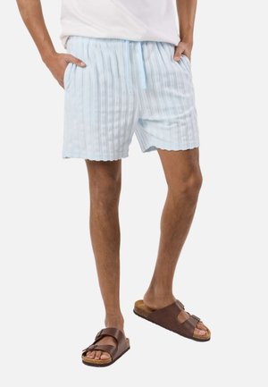 Mężczyzna noszący jasnoniebieskie, pasiaste spodenki z kieszeniami i brązowe sandały na podwójnych paskach, stojący z rękami w kieszeniach.