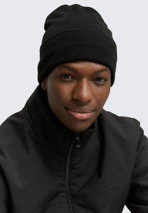 Zwarte geribbelde gebreide beanie met omgeslagen manchet. Draagt het met een zwarte zip-up jas, tegen een lichtgrijze achtergrond.