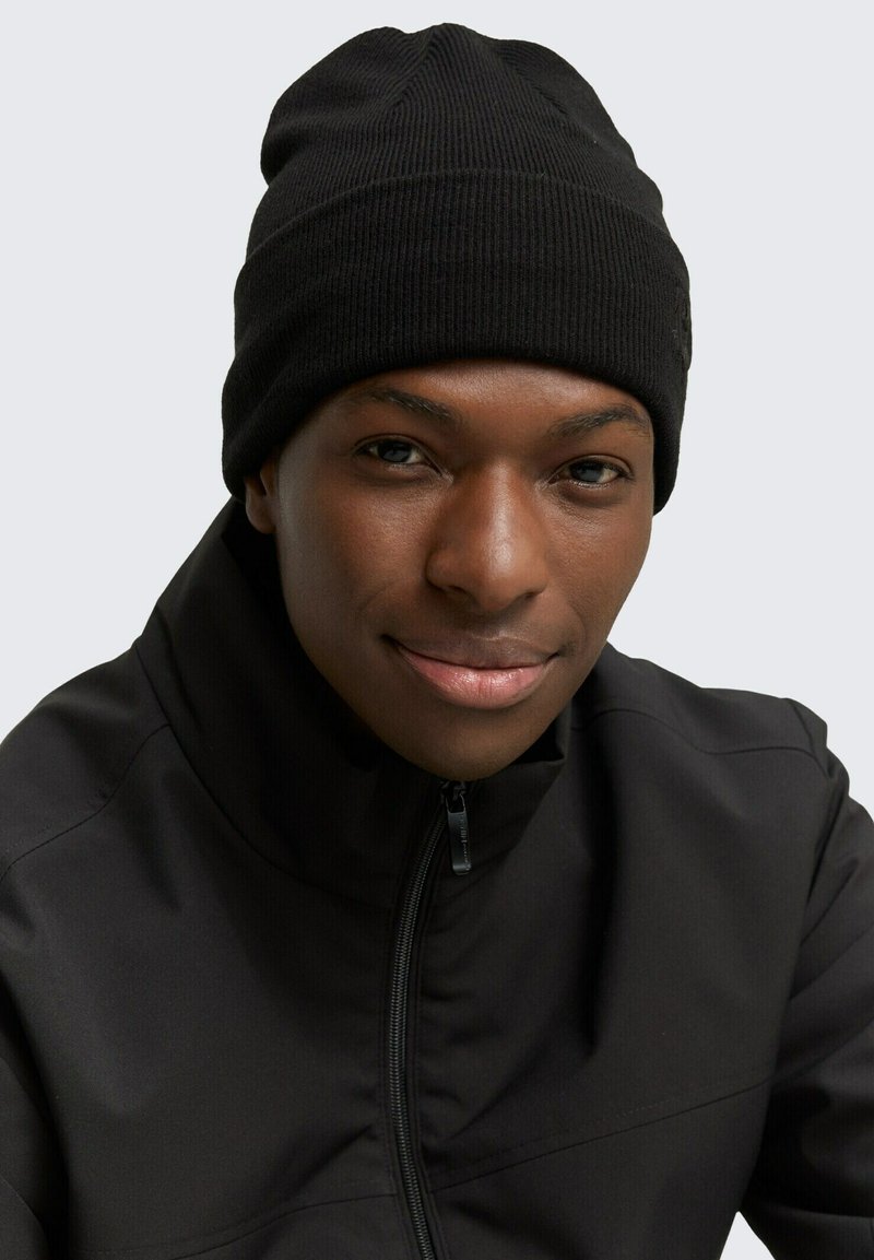 Căciulă neagră din tricot cu model canelat, cu margine pliată. Purtată cu o jachetă neagră cu fermoar, pe un fundal gri deschis.