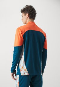 Maglia a maniche lunghe con design a blocchi di colore nei toni arancio, blu scuro e un pannello laterale a motivo in bianco, arancio e grigio.
