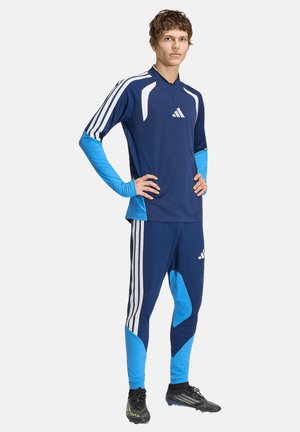 Joven de pie con las manos en las caderas, vistiendo ropa deportiva Adidas azul y azul claro con tres rayas blancas y tacos negros.
