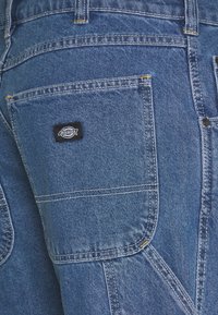 Blå denimbyxor med en ficka i lappa, gula sömmar som accentuerar, och en svart Dickies-logotag på bakfickan.