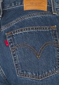 Poche arrière d'un jean en denim bleu avec une couture orange, une étiquette en cuir marron et une petite étiquette en tissu rouge sur le côté.