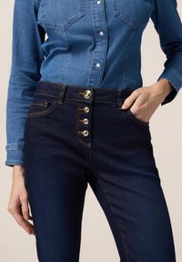 Los jeans de mezclilla azul oscuro presentan una cintura alta, cierre de cinco botones y costuras en contraste. La tela tiene una textura suave con un diseño clásico de bolsillos.