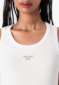Hvit ribbestrikket tanktopp med rund hals og et rektangulært logopatch med "JOOP! JEANS" i gull. Glatt tekstur, tettsittende design.