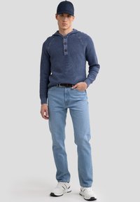 Tamsiai mėlynas riboto mezgimo džemperis su šnurku ir sagų užsegimu, derinamas su šviesiai mėlynomis džinsomis ir baltais sportiniais batais. Atsitiktinis aprangos derinys.