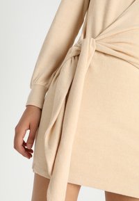 Robe portefeuille beige en tissu doux, avec un détail noué à la taille, des manches longues et une coupe droite qui tombe au-dessus du genou.