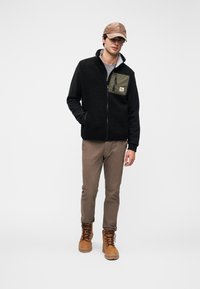 Μαύρο fleece μπουφάν με μια τσέπη με πράσινο φερμουάρ, γκρι πουκάμισο, καφέ παντελόνι και γήινα μποτάκια. Το μοντέλο φορά καφέ καπέλο, στέκεται μπροστά σε απλό φόντο.