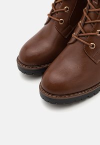 Brune læranklestøvler med afrundede tæer, struktureret overdel, snøre design, guldfarvet øjet hardware og robuste sorte såler.