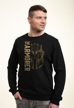 Jeune homme aux cheveux courts portant un sweat-shirt noir avec le texte doré « The Armorer » et un motif de casque de style Mandalorien, regardant vers la droite.