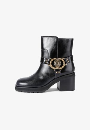 Zwarte leren enkelboot met een chunky hak, voorzien van een decoratieve gouden gesp en metalen studs op een band. Blessure textuur en elegante uitstraling.