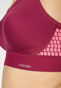 Burgunderfarget sports-bh med jevn tekstur, med et rosa geometrisk mønster på siden og en logo på båndet. Designet for komfort og støtte.