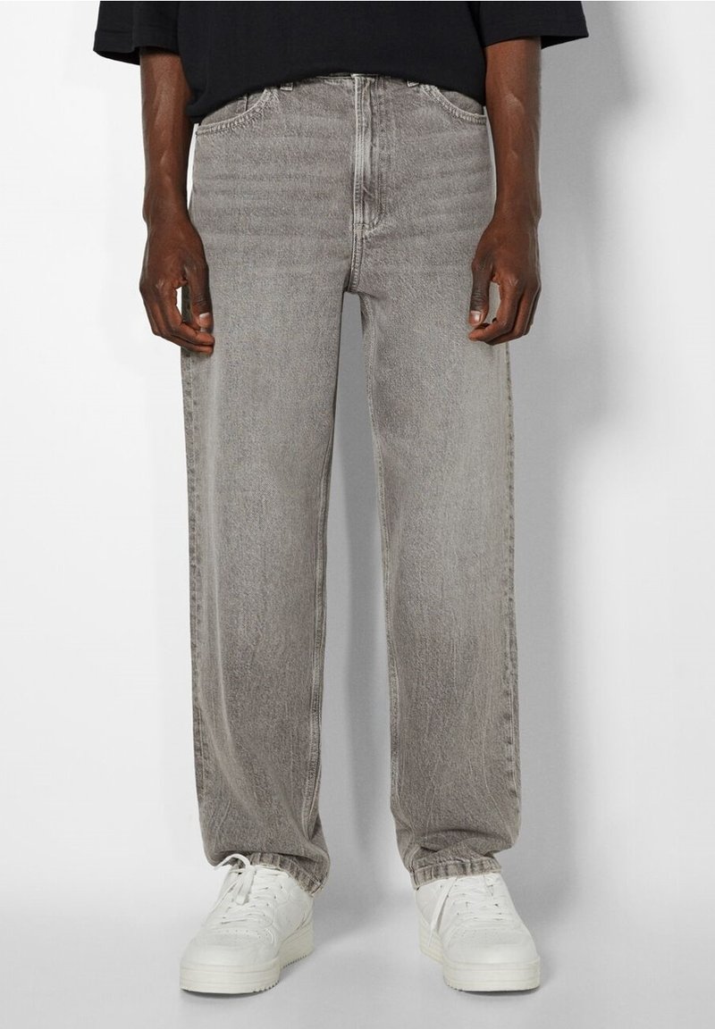 Homme portant un jean droit gris délavé et des baskets blanches, debout les mains détendues sur les côtés devant un fond uni.