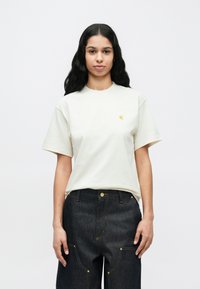 Mujer con el cabello largo y negro, vistiendo una camiseta de manga corta color blanco hueso y jeans oscuros de tiro alto, de frente contra un fondo liso.