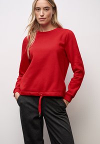 Roter Baumwollpullover mit Rundhalsausschnitt, gerippten Bündchen und verstellbarem Kordelzug am Saum. Kombiniert mit dunkelgrauen Stoffhosen.