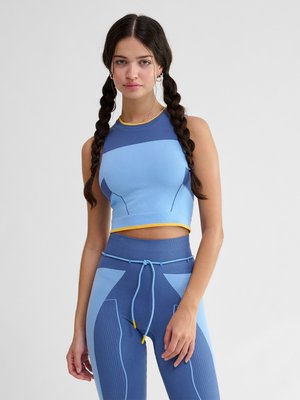 Ung kvinde med mørkt fletning hår bærer blå og lyseblå atletisk crop top og leggings med gule accenter, stående mod en ensfarvet baggrund.