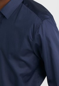 Marineblauwe shirt met een puntige kraag, een gladde textuur en lange mouwen; heeft een donkere, glanzende stof op de schouder.