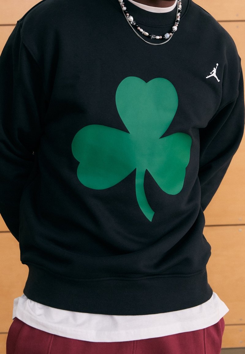 Personne portant un sweat-shirt noir avec un grand trèfle vert et un petit logo Jumpman blanc, superposé à une chemise blanche et un pantalon rouge.