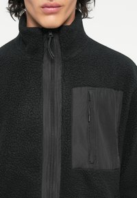 Schwarze Fleecejacke mit hohem Kragen, Reißverschluss und strukturiertem Körper. Enthält eine glatte schwarze Brusttasche aus Stoff mit Reißverschluss.
