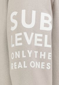 Helles graues Textil mit auffälliger weißer Schrift, die "SUB LEVEL ONLY THE REAL ONES" sagt. Der Stoff sieht weich und gestrickt aus.